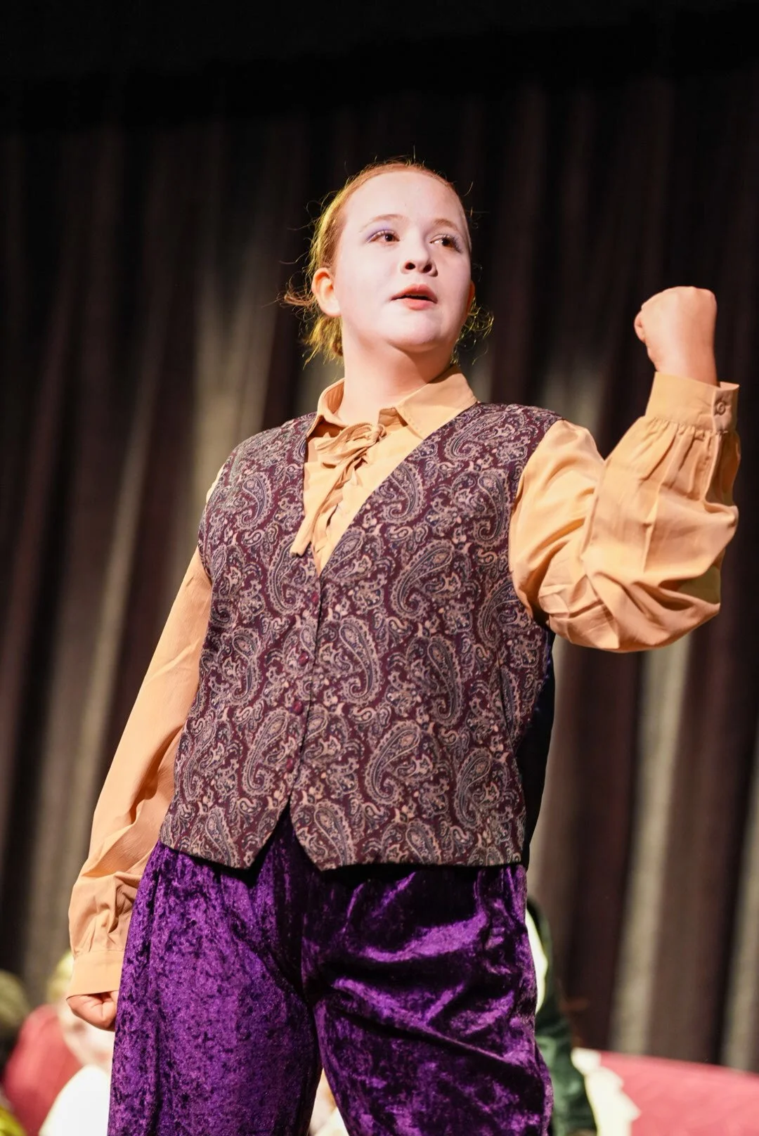 A young girl on stage with a confident expression, wearing a vintage-style vest, a beige shirt with puffy sleeves, and purple velvet pants, raising her arm in a gesture of strength or determination.