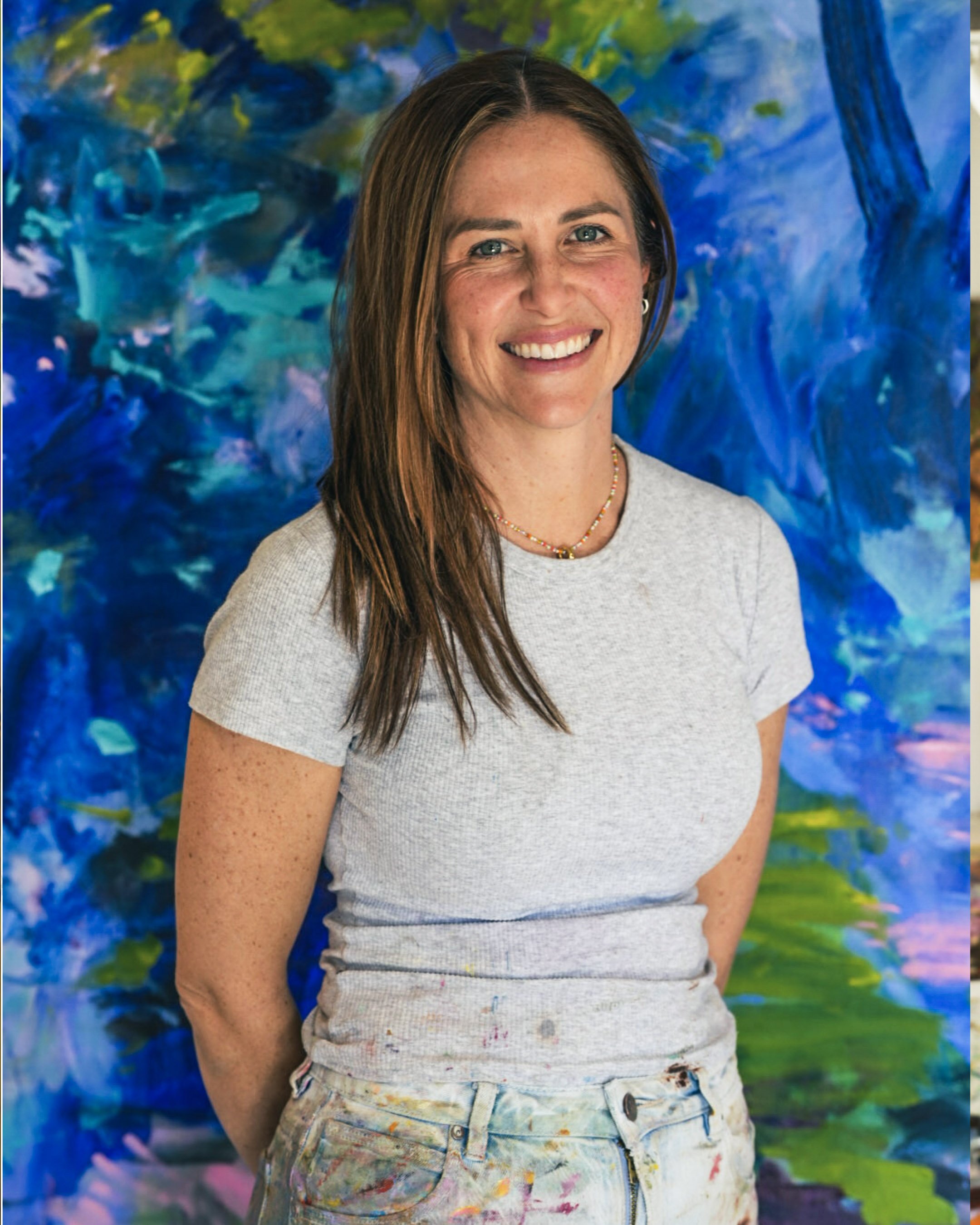 A woman with long brown hair, smiling, wearing a gray t-shirt and multicolored painted jeans, standing in front of an abstract blue and green painted background.