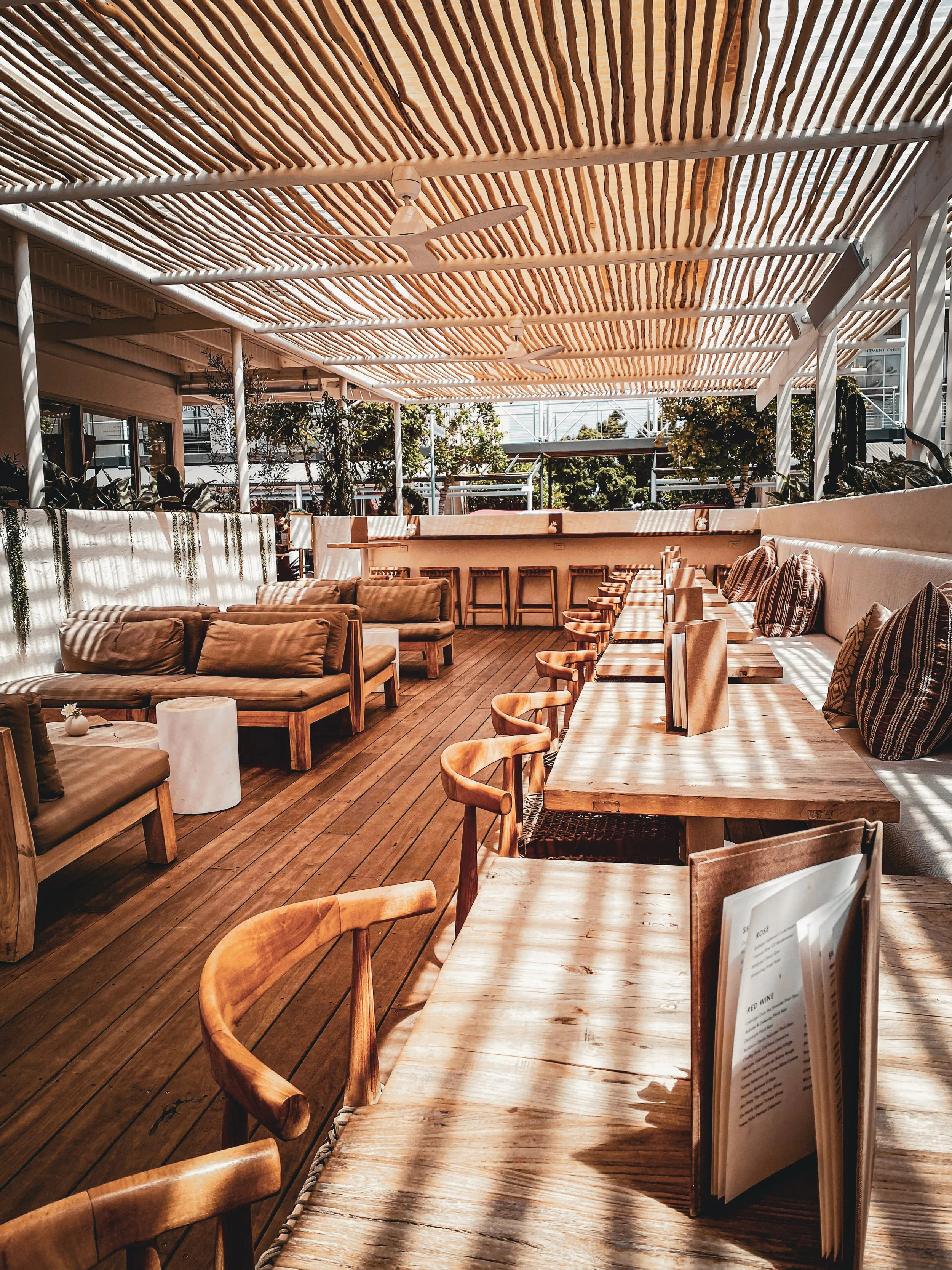 Outdoor rooftop bar with wooden furniture, tables with menus, cushioned benches, tables and chairs, ceiling with wooden slats, ceiling fans, and a railing with greenery.