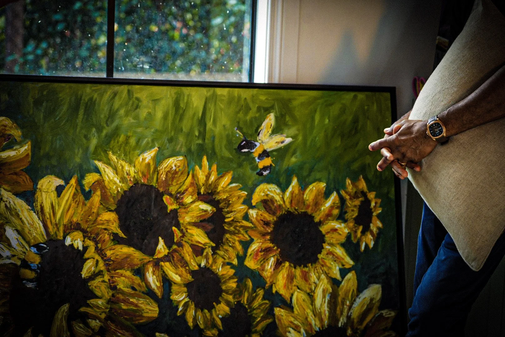 Painting of sunflowers with a bee hovering above, displayed in front of a window with greenery outside.