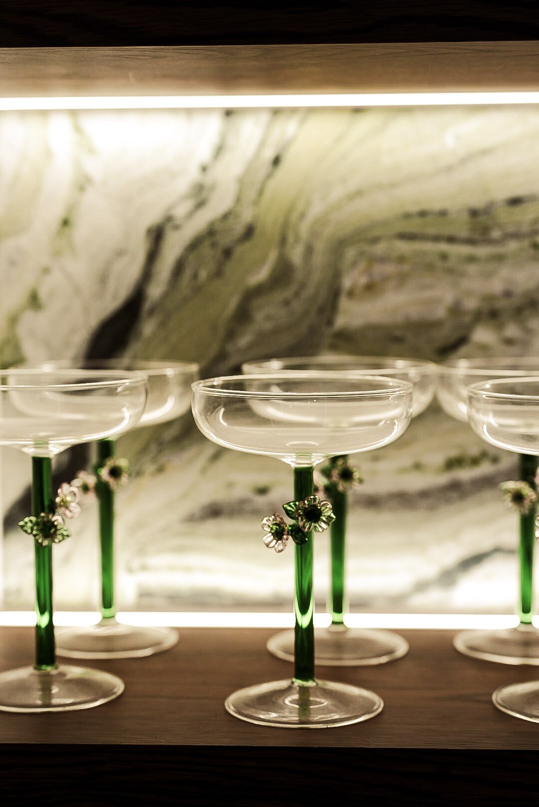 Multiple cocktail glasses with green stems and decorative flowers, arranged on a wooden surface with a marbled wall in the background.