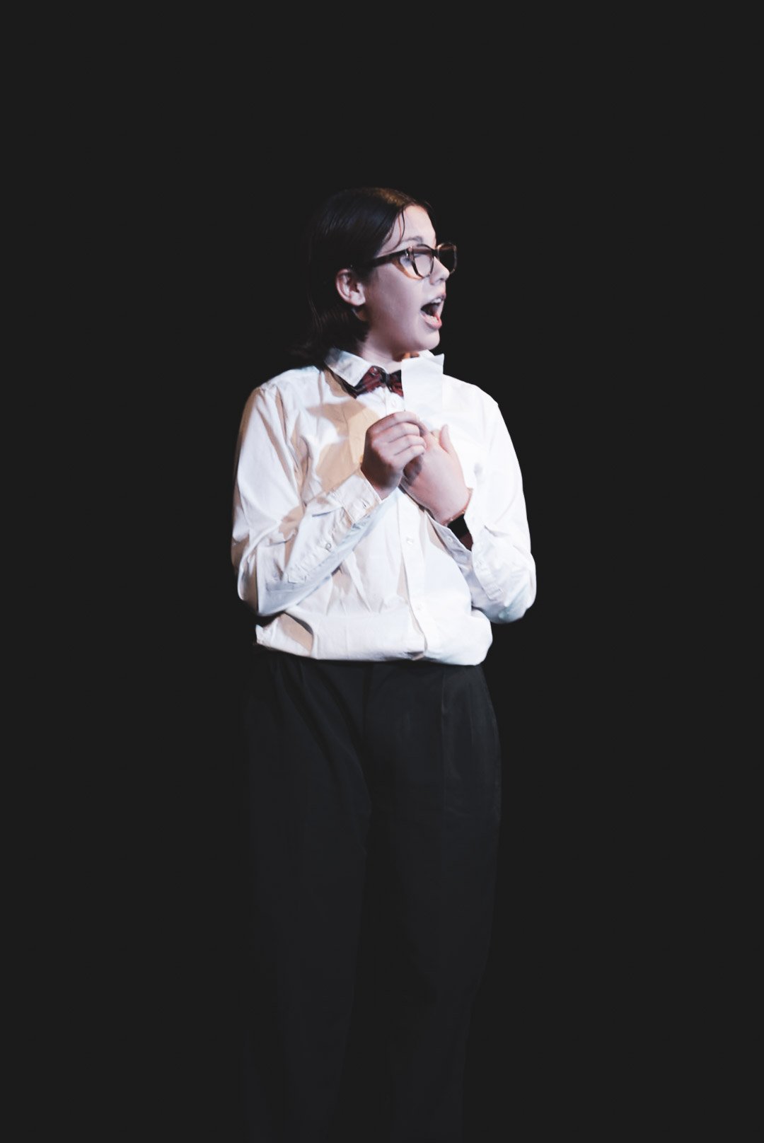 A young woman with glasses and dark hair, dressed in a white shirt and black pants, standing on a black stage, appearing to speak or sing, holding a small object with an emotional expression.