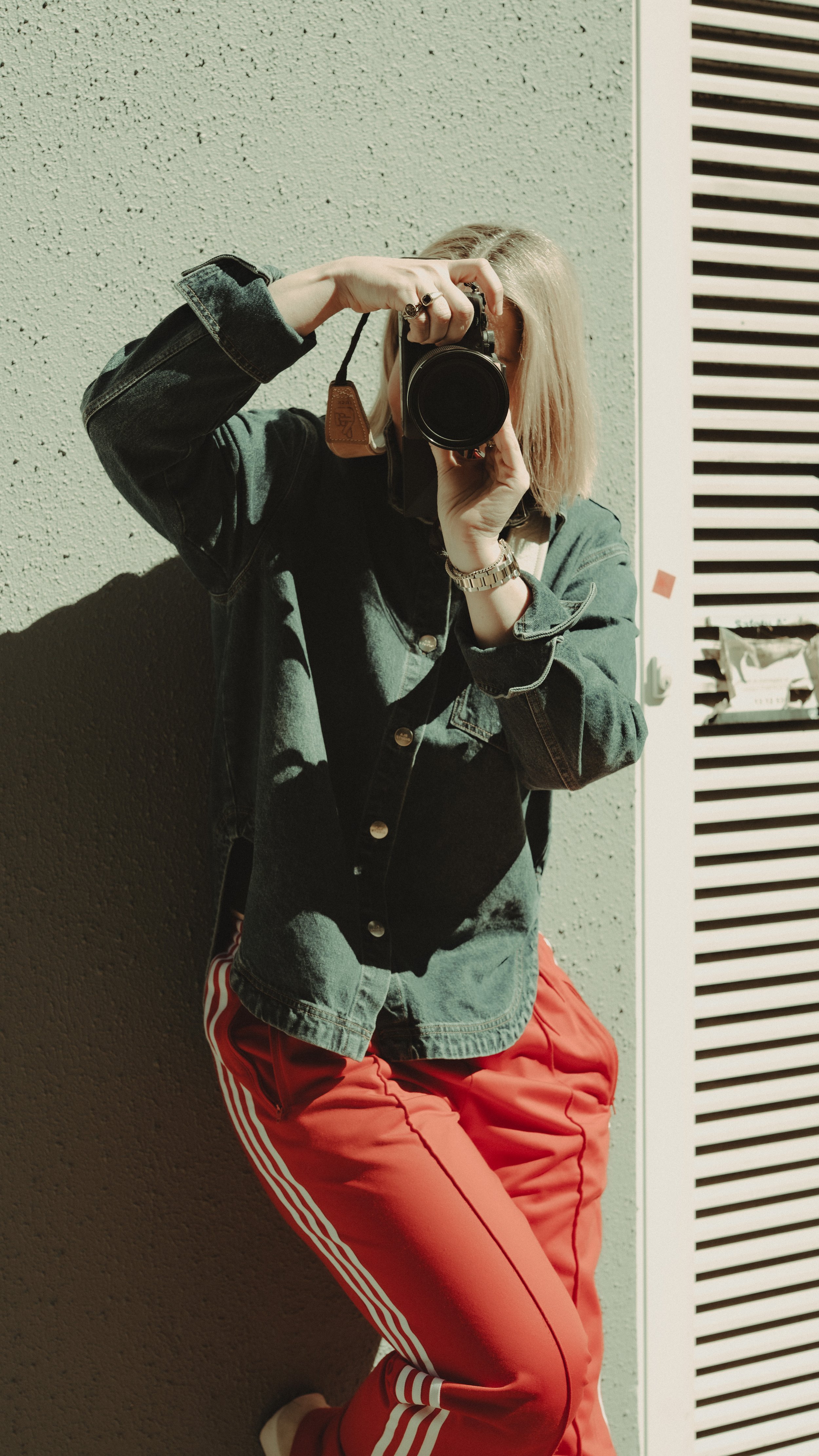 Person taking a selfie with a camera, standing against a textured wall with a radiator nearby. They are wearing a black denim jacket and red Adidas track pants.