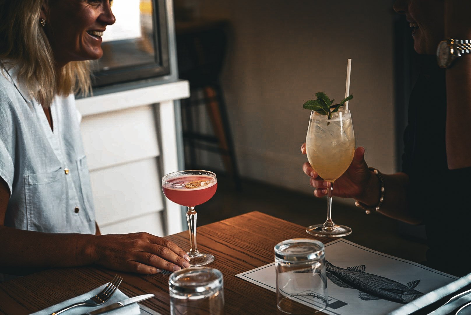 Two women at a restaurant, smiling and holding cocktails; one with a pink drink and the other with a yellow drink garnished with mint and a straw.