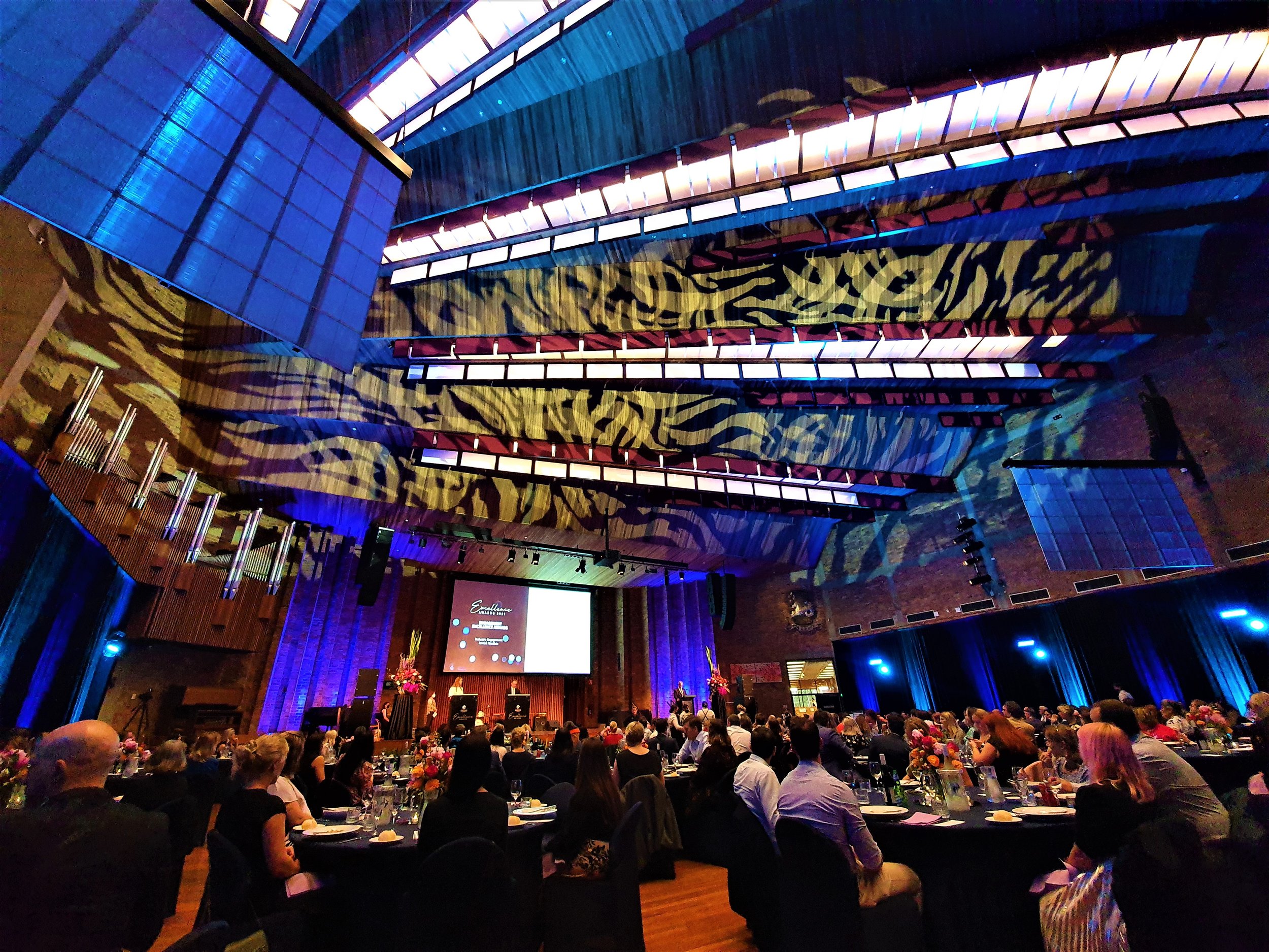 Indoor event in a theater with attendees seated at tables. The ceiling displays colorful abstract lighting patterns. A stage is visible with a large screen and flowers. People engaged in an event or presentation.