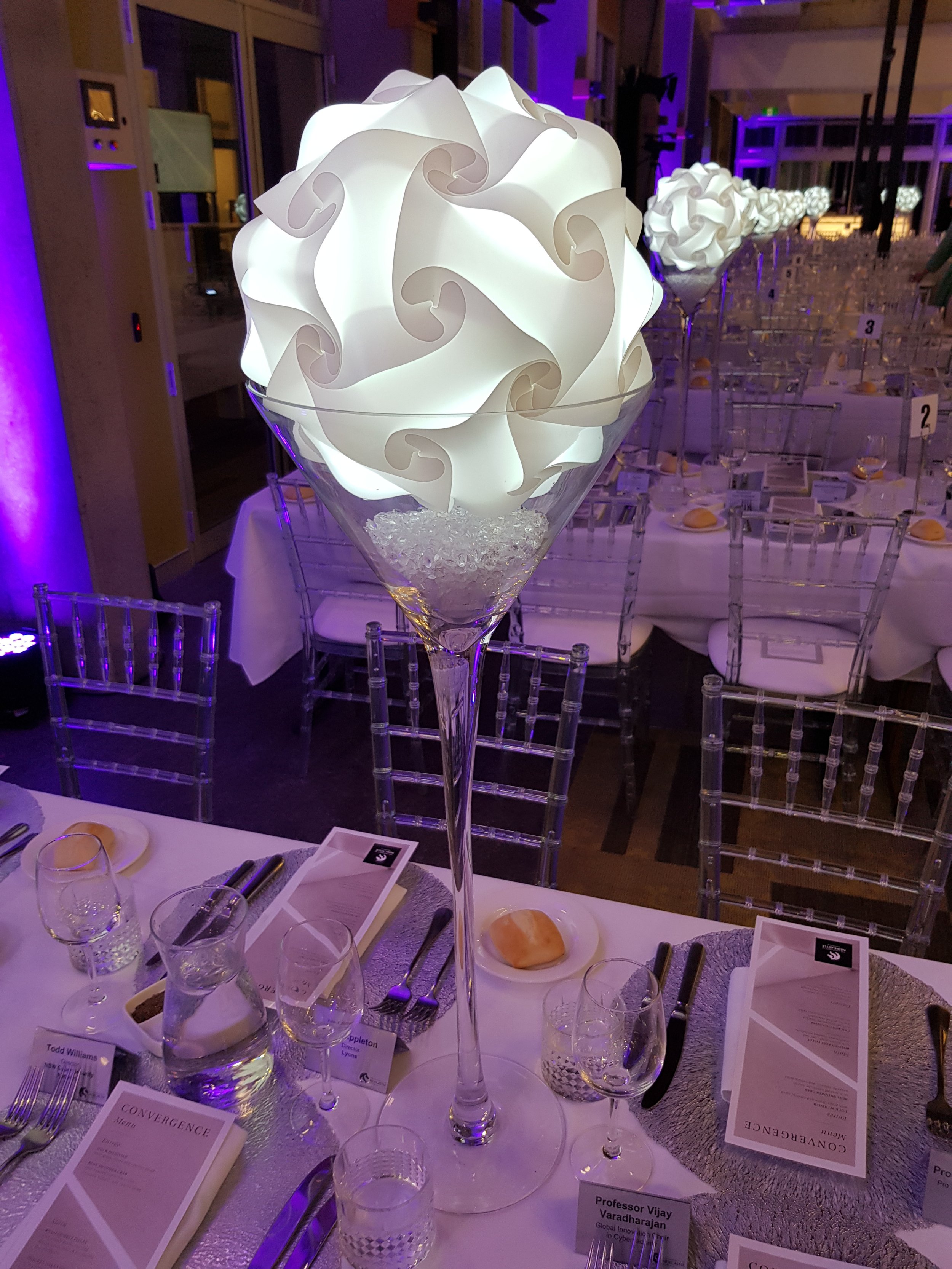 Decorative table setup with a tall glass centerpiece containing a glowing, intricate spherical lamp made of white geometric shapes. The table is set with plates, menus, and glassware, surrounded by transparent chairs. Purple lighting creates an elega