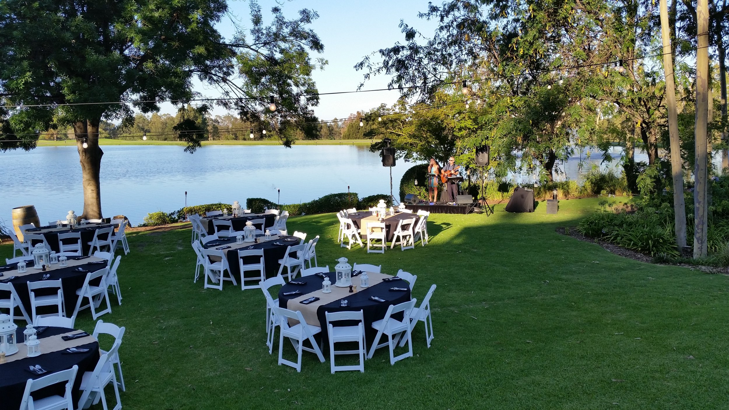 Outdoor event setup with round tables covered in black and white linens, white chairs, a stage with musicians, green lawn, trees, and a lake in the background.