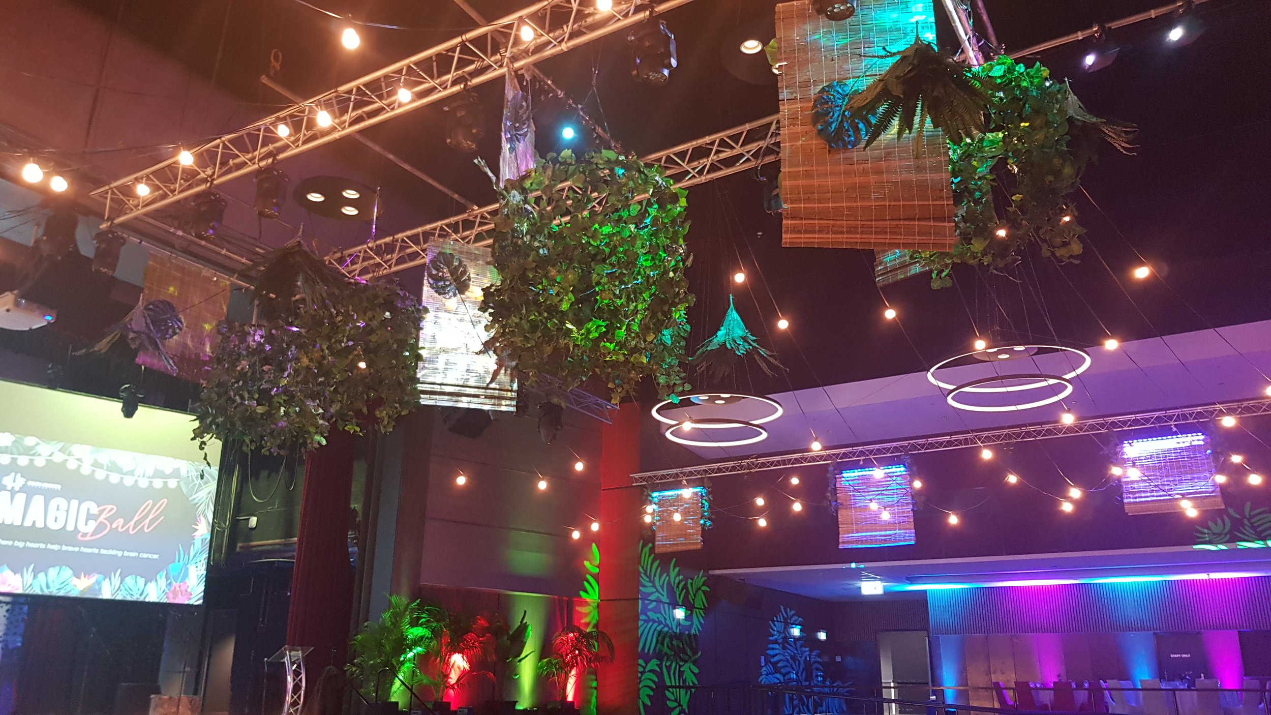 Decorated indoor venue with hanging greenery, string lights, and ring lights. A screen displays 'Magic Ball' event details with colorful stage lighting and tropical plants.