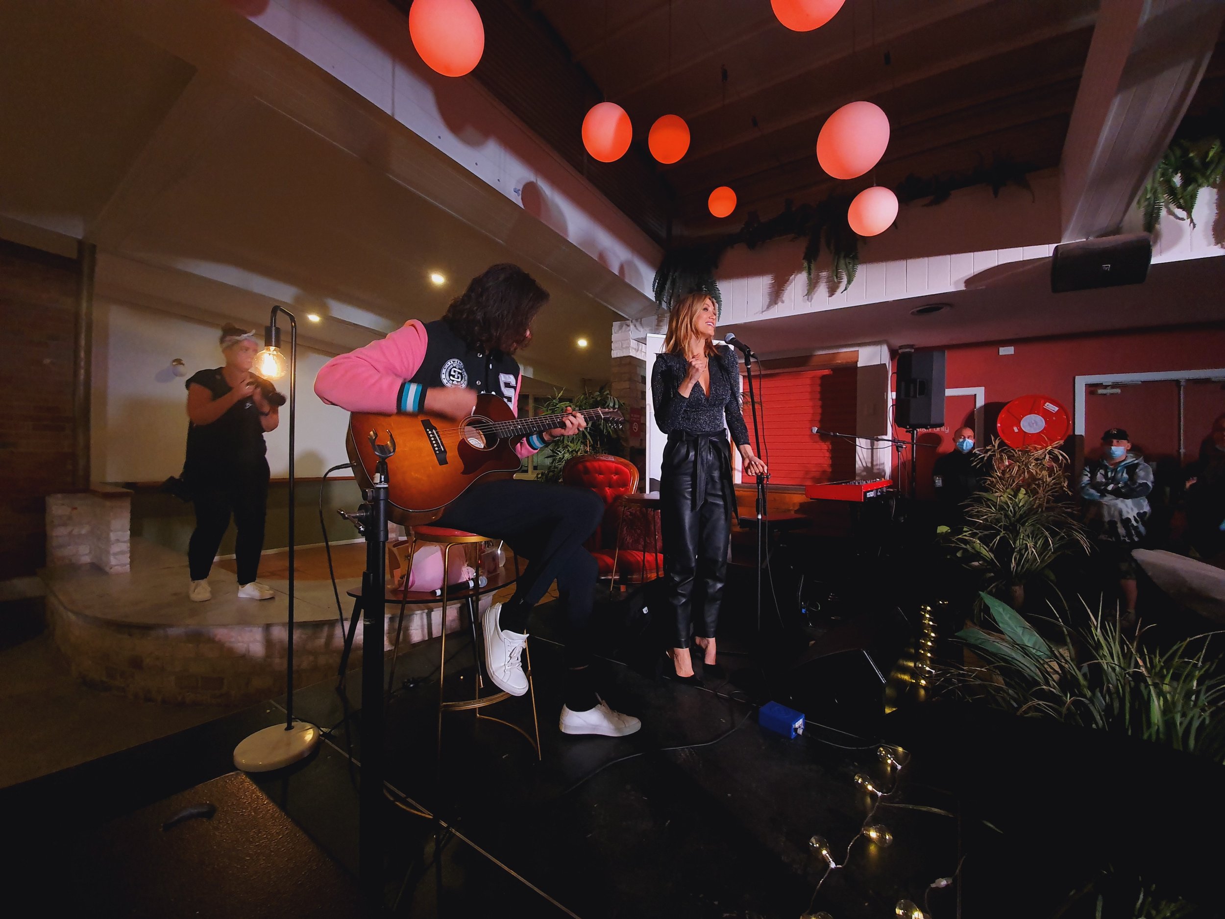 Woman singing on stage with guitarist and photographer, dimly lit venue, red hanging lights, audience watching.