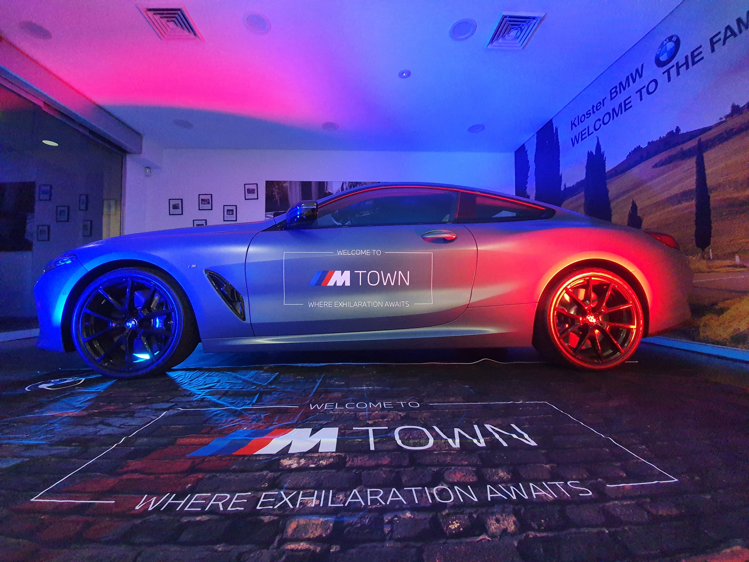 BMW coupe car displayed in a showroom with blue and red lighting, featuring 'M Town' branding and signage on the floor, and a wall poster that says 'Kloster BMW Welcome to the Family.'