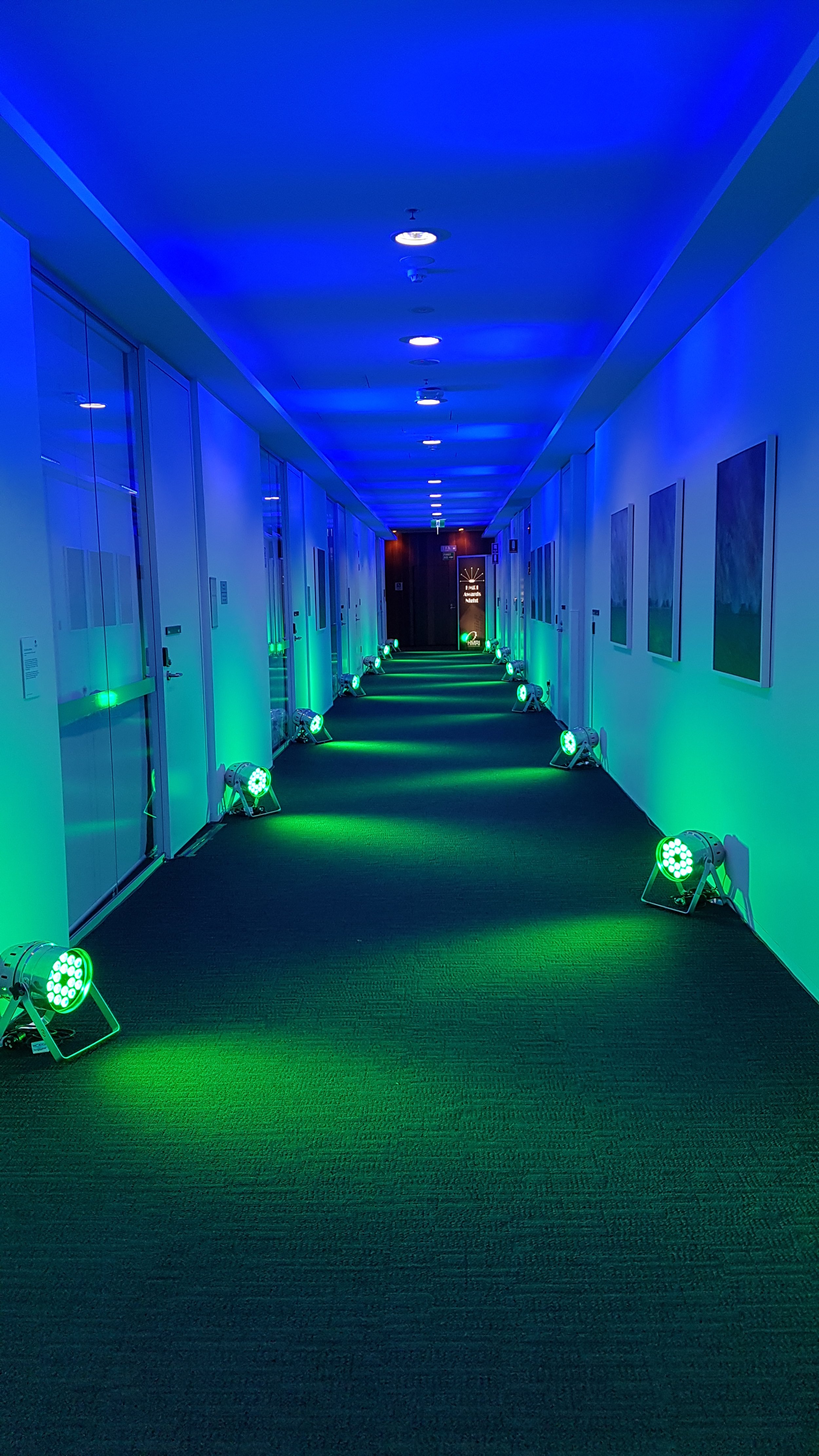 A corridor illuminated with blue and green lights creating a vibrant ambiance. The walls have framed art pieces, and spotlights are arranged along the floor.