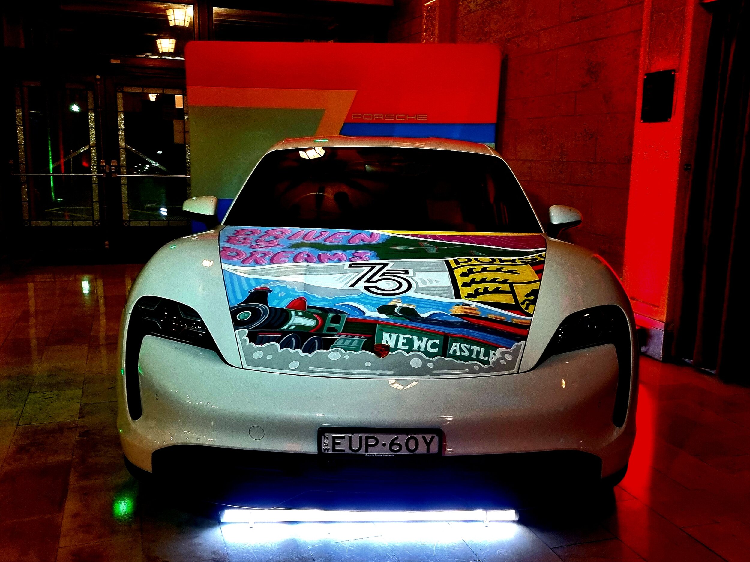 White Porsche car with colorful artwork on the hood in a dimly lit area, featuring a train and text "Driven by Dreams" and "75". Porsche logo at the back.