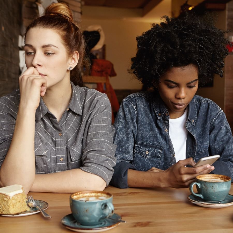 Two women sitting at a wooden table in a cozy cafe, with cups of coffee and a slice of cake, one looking thoughtful and the other looking at her phone.