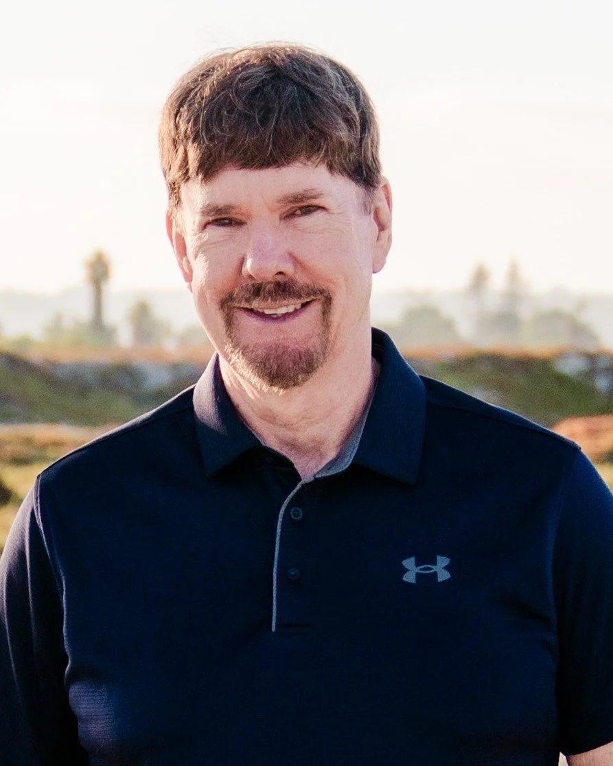 Man smiling outdoors in a dark blue Under Armour polo shirt, with a background of a sunny landscape with trees.