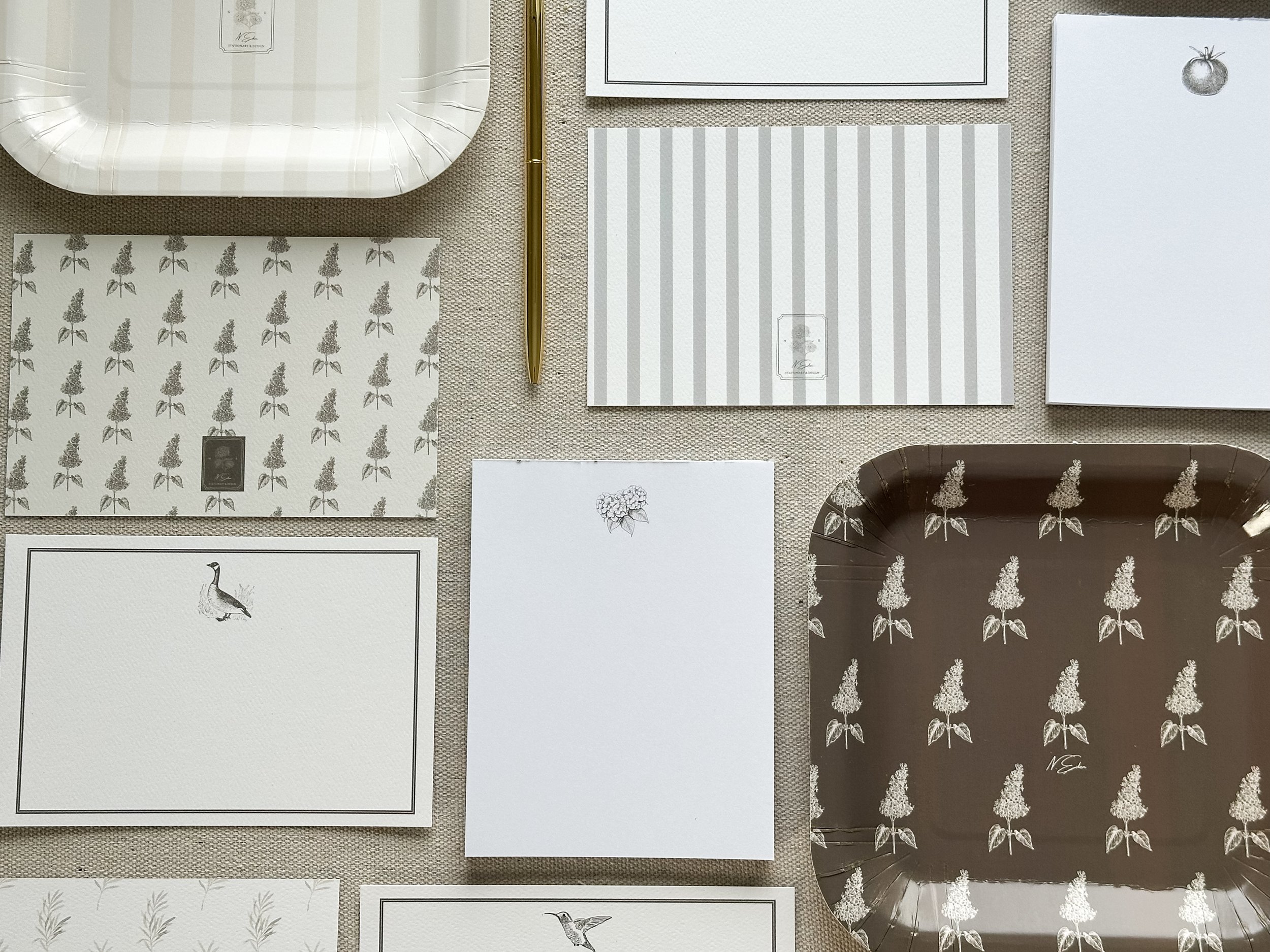 Flat lay of various stationery items including notecards with bird, floral, and striped designs, a gold pen, a white tray, and a brown tray, all arranged on a beige fabric surface.