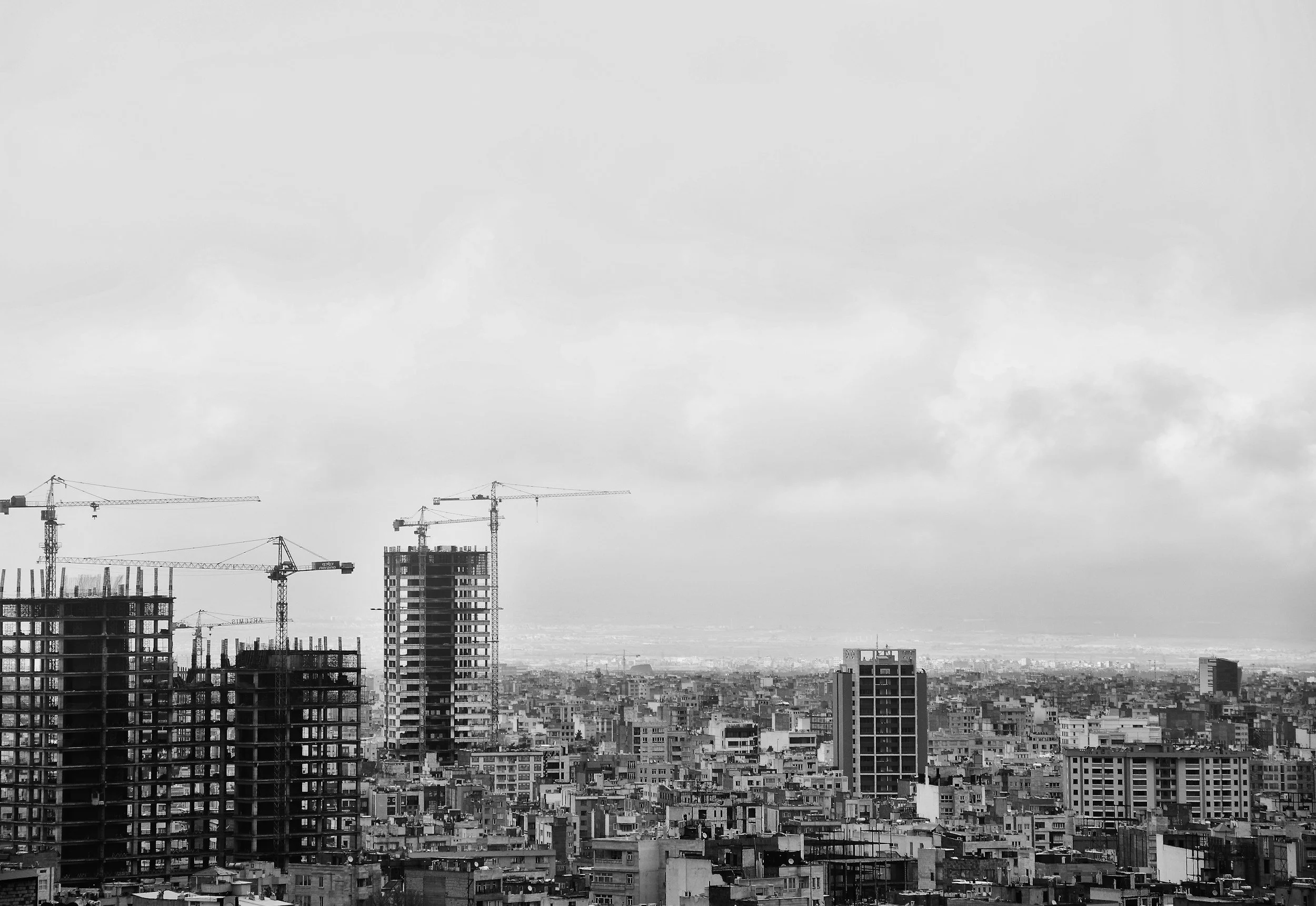 Black and white cityscape with construction cranes and high-rise buildings under construction against a cloudy sky.