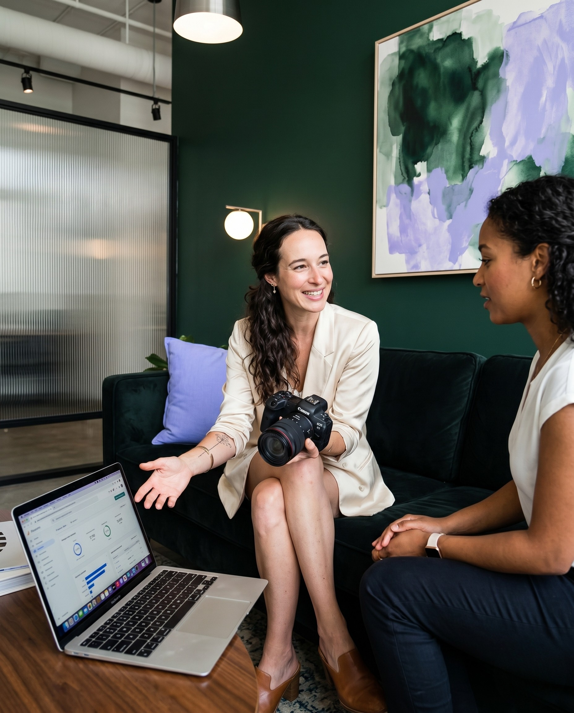 Two women are sitting on a dark sofa conversation in a modern office space, with one woman holding a camera. A laptop displaying a financial dashboard is on a coffee table nearby. The background features a green wall with abstract artwork, exposed ceiling pipes, and modern lighting.