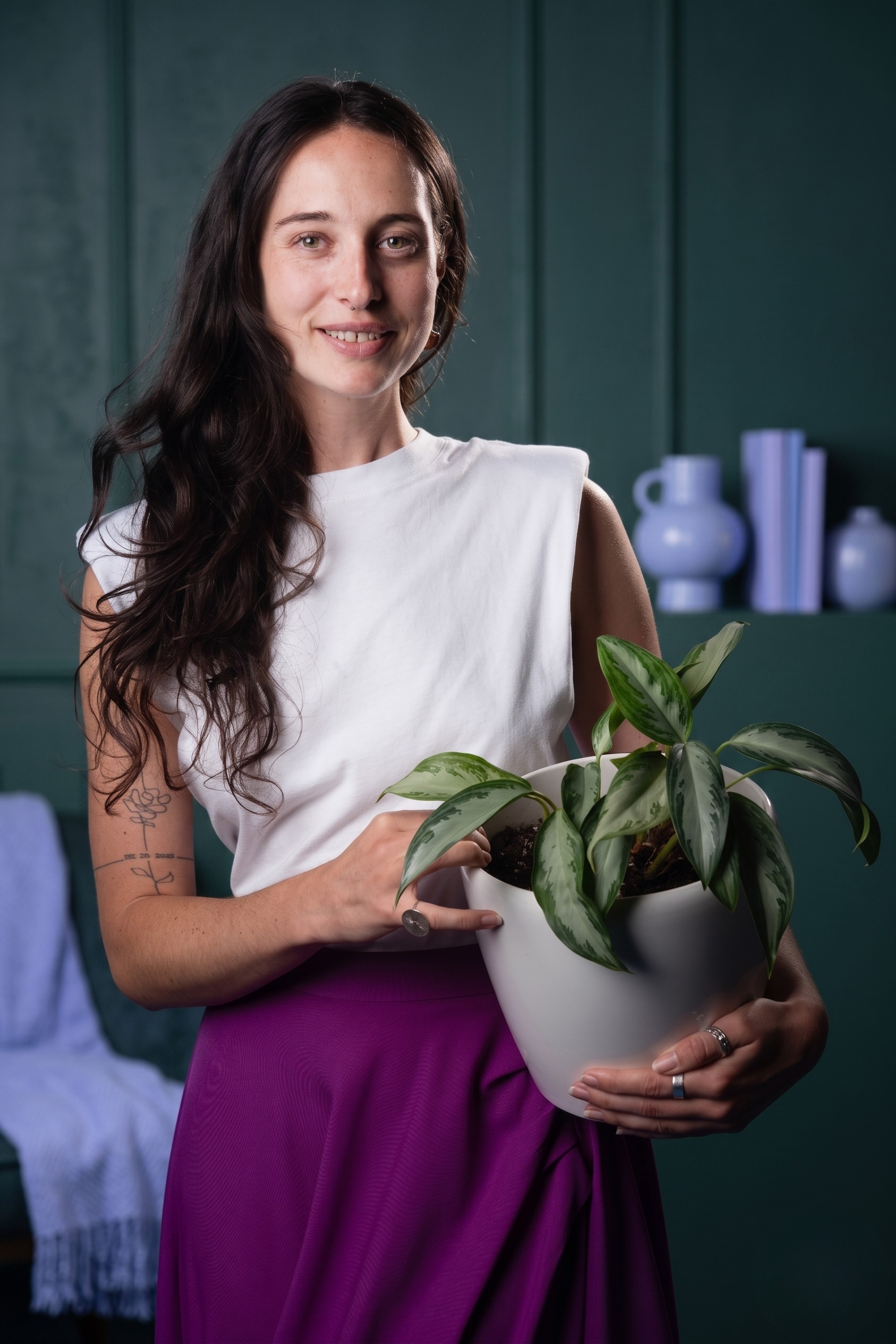 A young woman with long dark hair holding a potted plant with green and white leaves, smiling at the camera, with a teal background and decorative vases on a shelf behind her.
