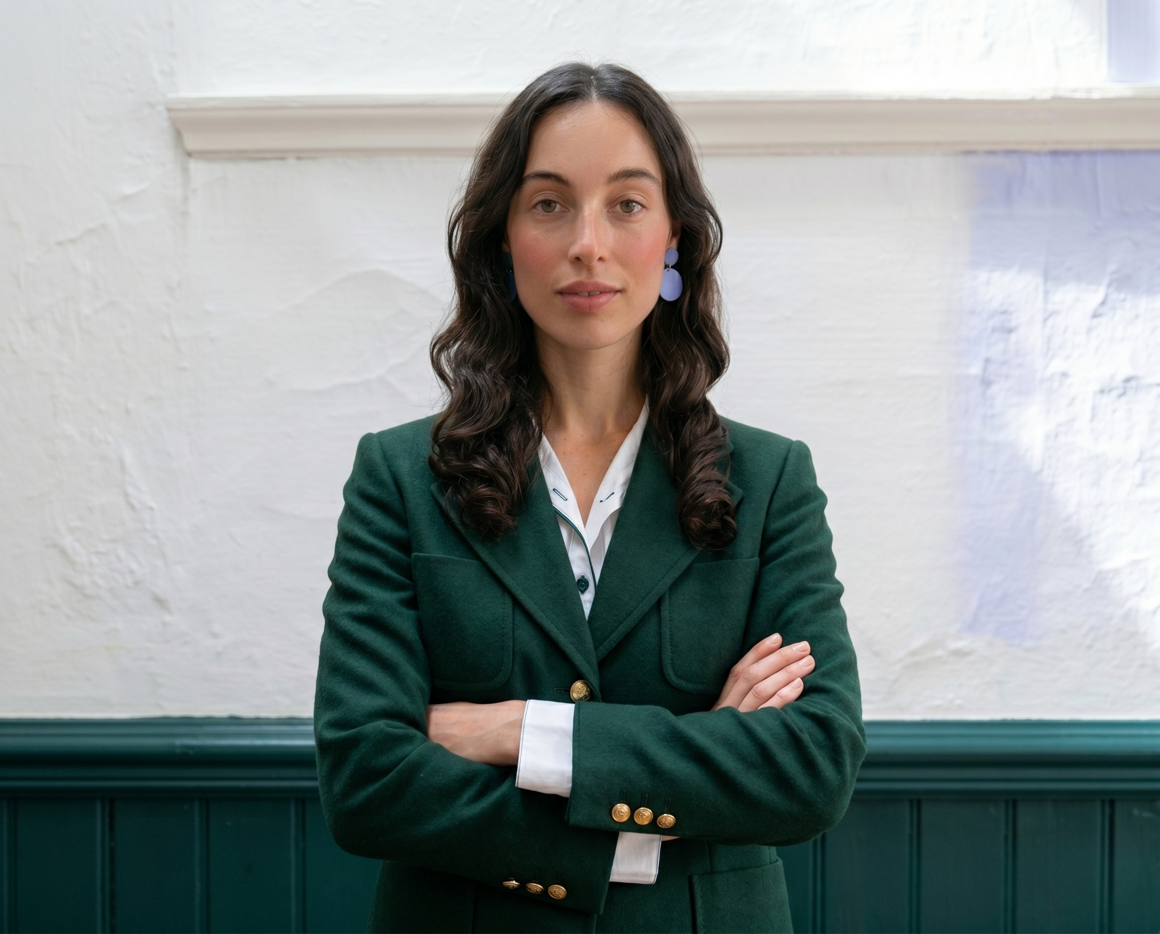 A woman with long, wavy dark hair and light skin standing with her arms crossed in front of a white textured wall and dark green paneling. She is wearing a dark green blazer with gold buttons over a white shirt.