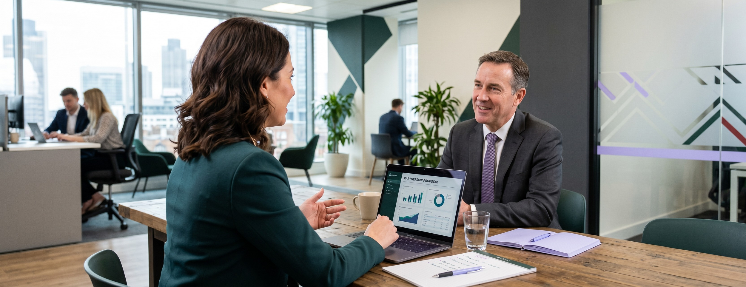 A woman and a man having a business meeting in a modern office with large windows. The woman is showing a digital tablet displaying charts to the man, who is smiling. In the background, other people are working at their desks.
