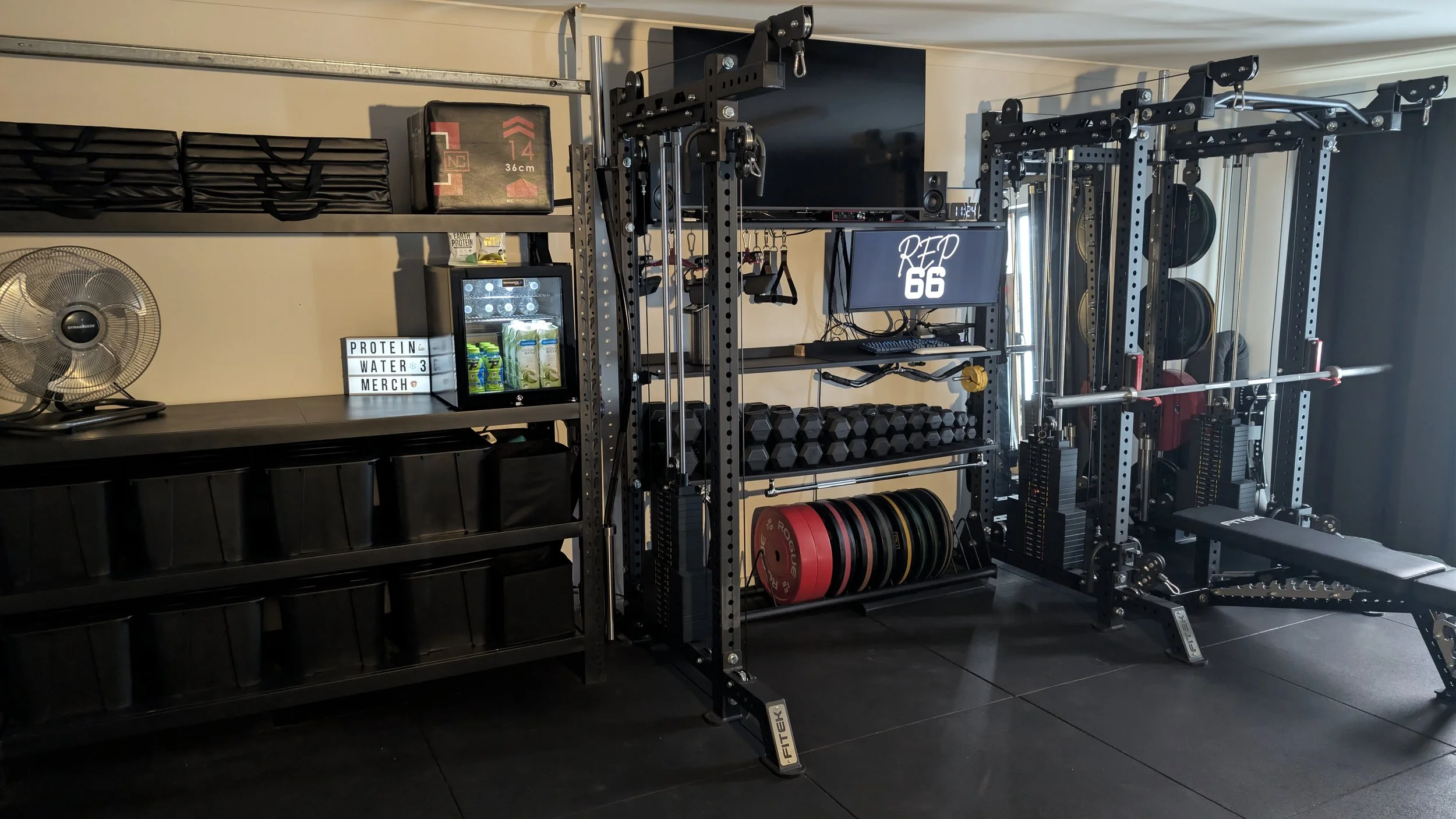 Home gym with weightlifting equipment, shelves with black storage bins, a fan, a small fridge with drinks, and a digital sign displaying 'PROTEIN WATER 3 MERCH'.