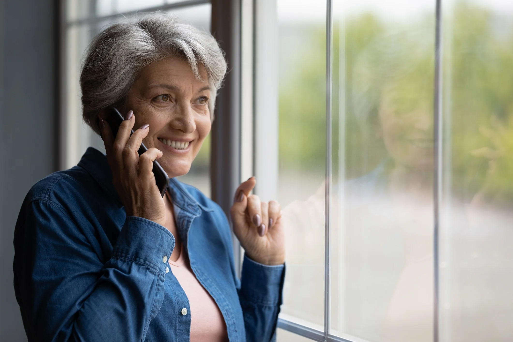 Smiling woman with gray hair talking on a cell phone while looking out a window
