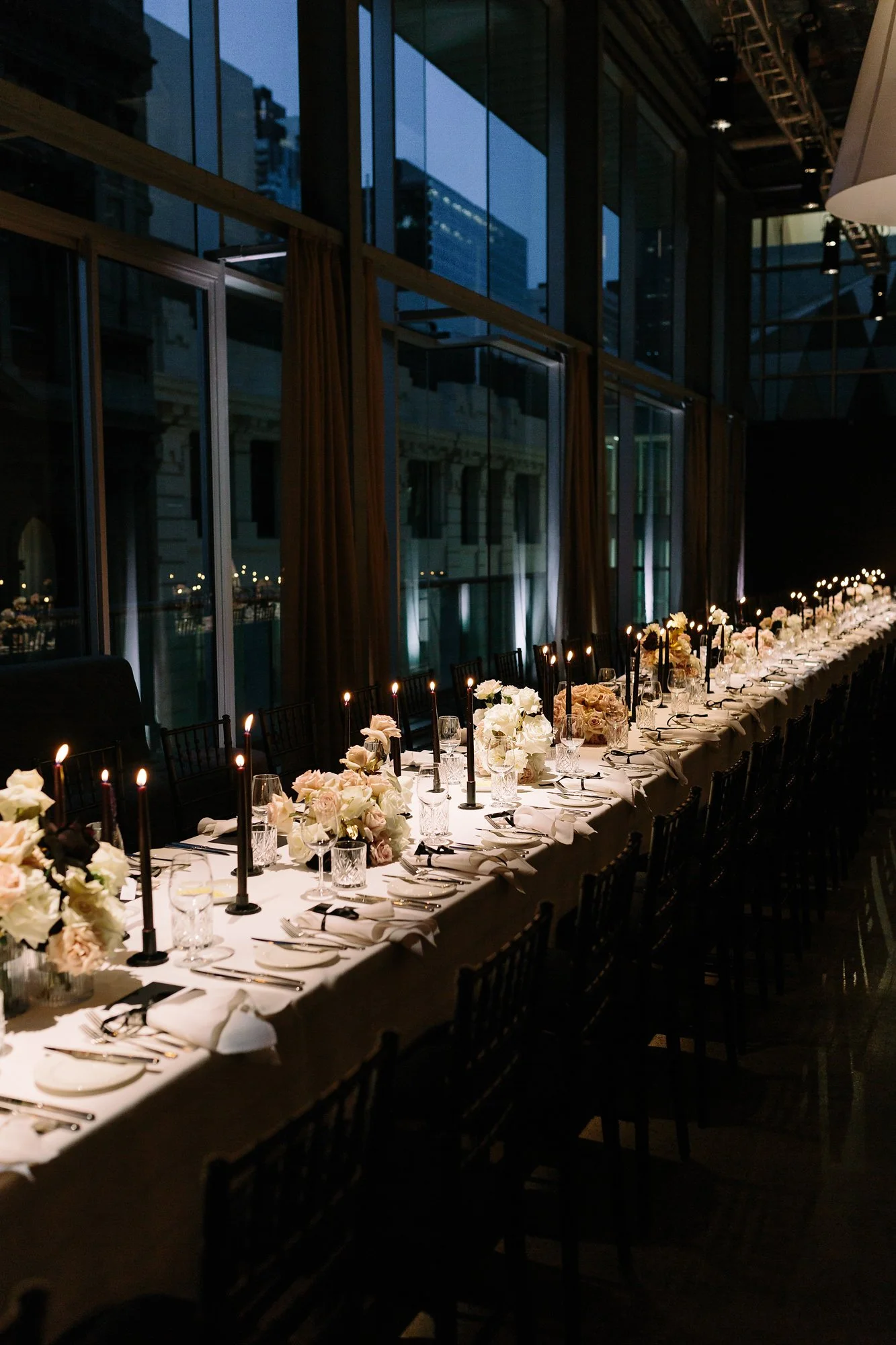 A long dining table set for a formal event with white tablecloths, floral centerpieces, black candles, glassware, and silver utensils. The table is indoors with large windows and city buildings visible outside at dusk.