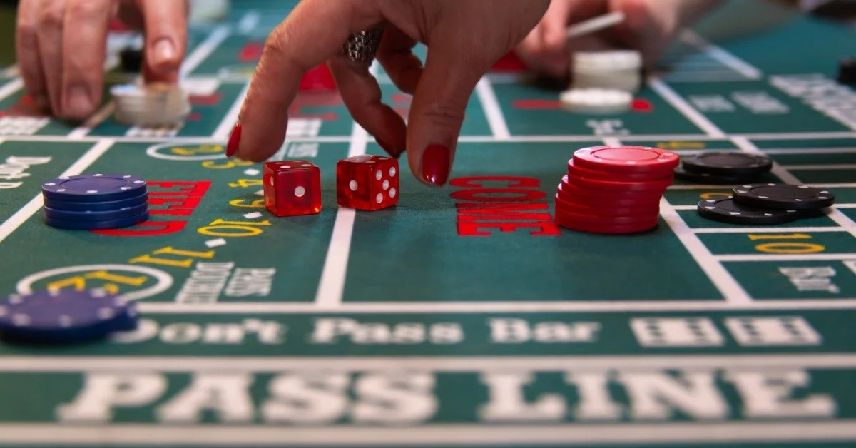 A hand reaches for red dice on a green casino table. Stacks of red, black, and blue chips sit on marked betting areas.