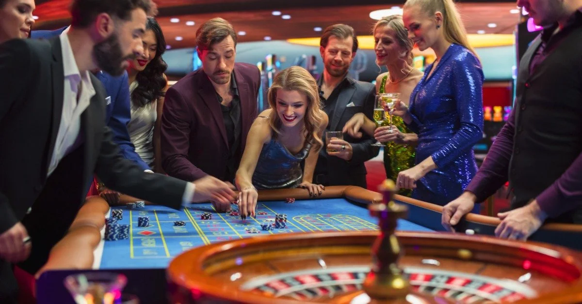 Adults in formal attire play at a casino table covered with chips, game markings, and a roulette wheel.