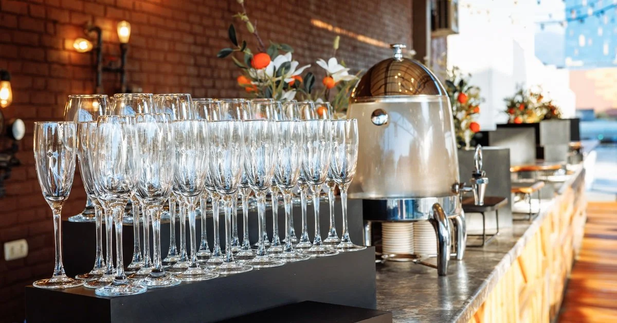 Champagne flutes are stacked in organized rows on a long buffet counter. A drink dispenser and dishes are further down.