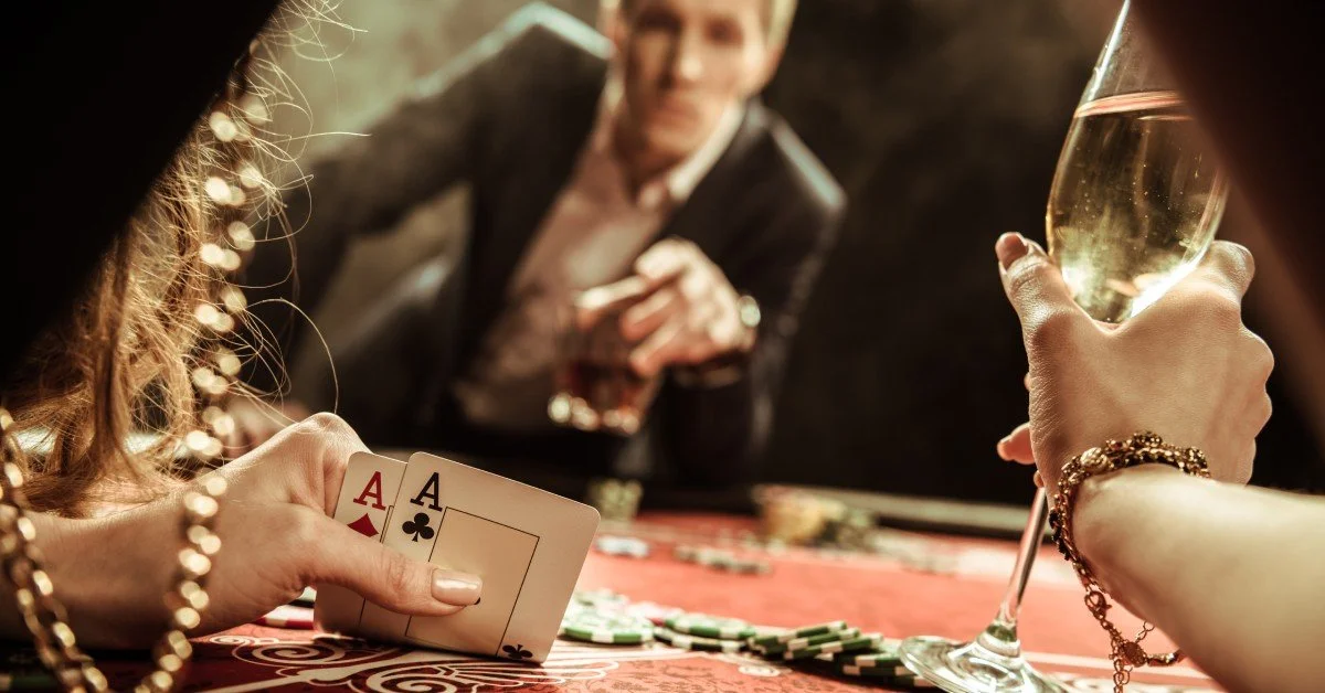 A woman looks at the two cards in her hand, an ace of diamonds and an ace of spades. The man across from her watches.