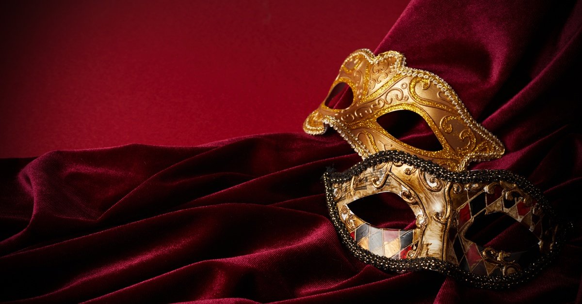 Two ornate masquerade eye masks lie on red velvet fabric. One is a gold color, while the other is gold, black, and red.
