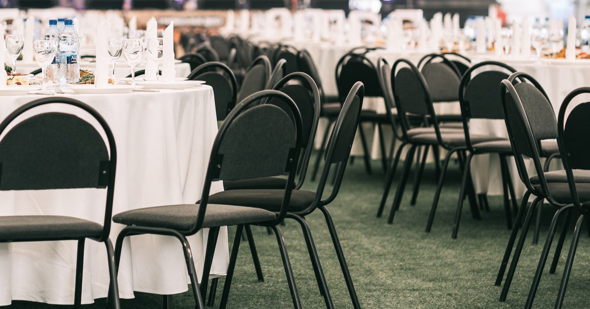Round tables with white tablecloths are surrounded by chairs in a large event space. Water bottles are on the tables.
