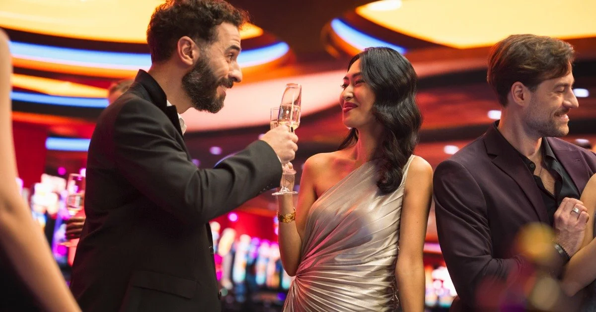 A man in a suit raises a champagne flute toward a woman wearing a silver dress. A man near them talks to someone else.