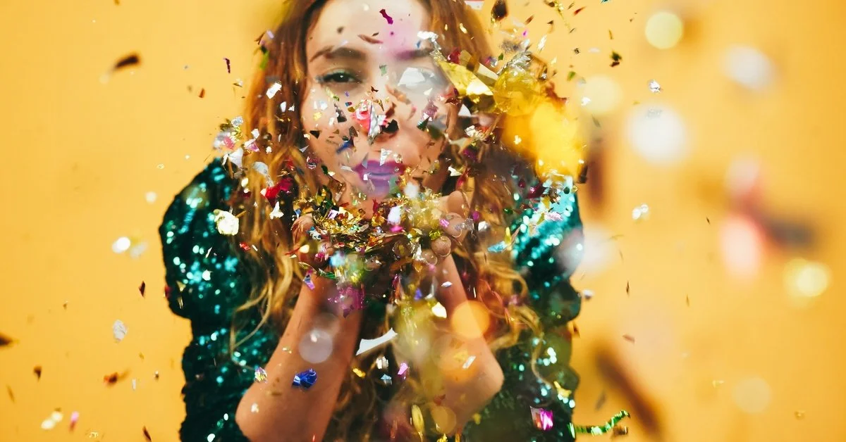 A woman in a green sequined top stands against a bright yellow background. She blows metallic confetti into the air.