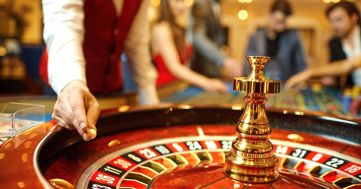A casino dealer holds a roulette ball above a shining roulette wheel. Players stand around the table in the background.
