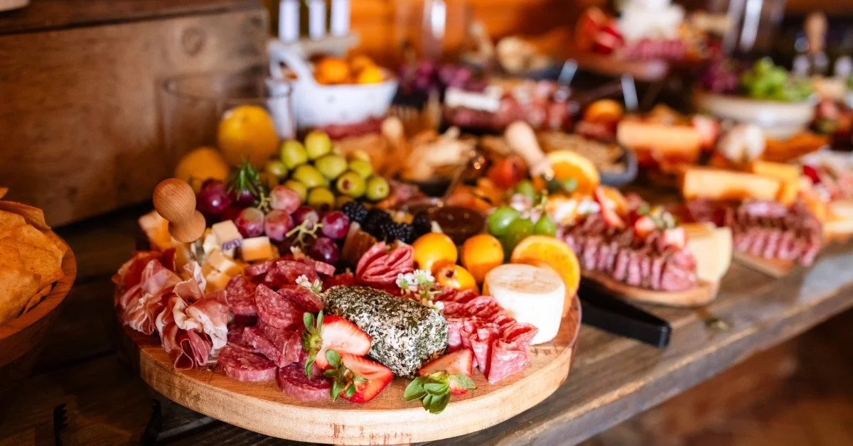 A grazing table features round boards topped with salami, prosciutto, cheese, strawberries, grapes, olives, and oranges.