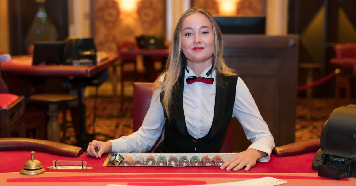 A slightly smiling casino dealer sits at a gaming table with stacks of multicolored chips and playing cards before her.