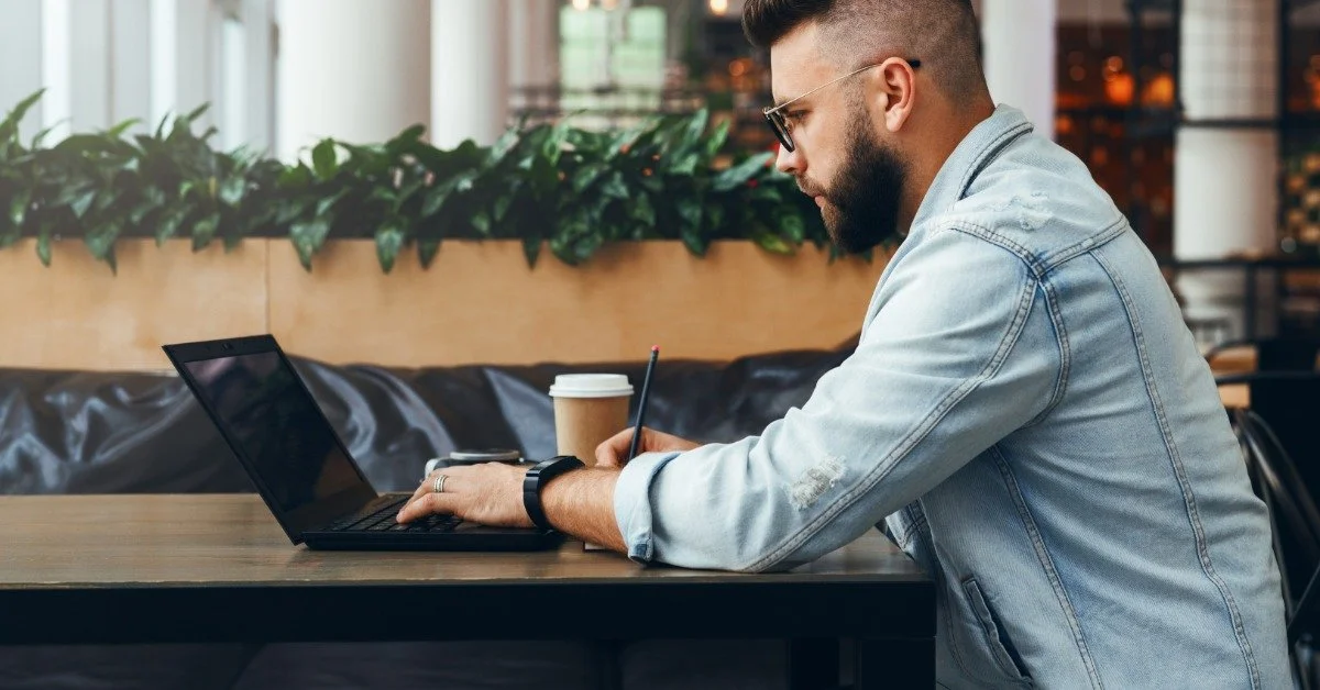 Side view of a man at a table with a laptop and a cup of coffee. He writes something as he looks at his laptop screen.