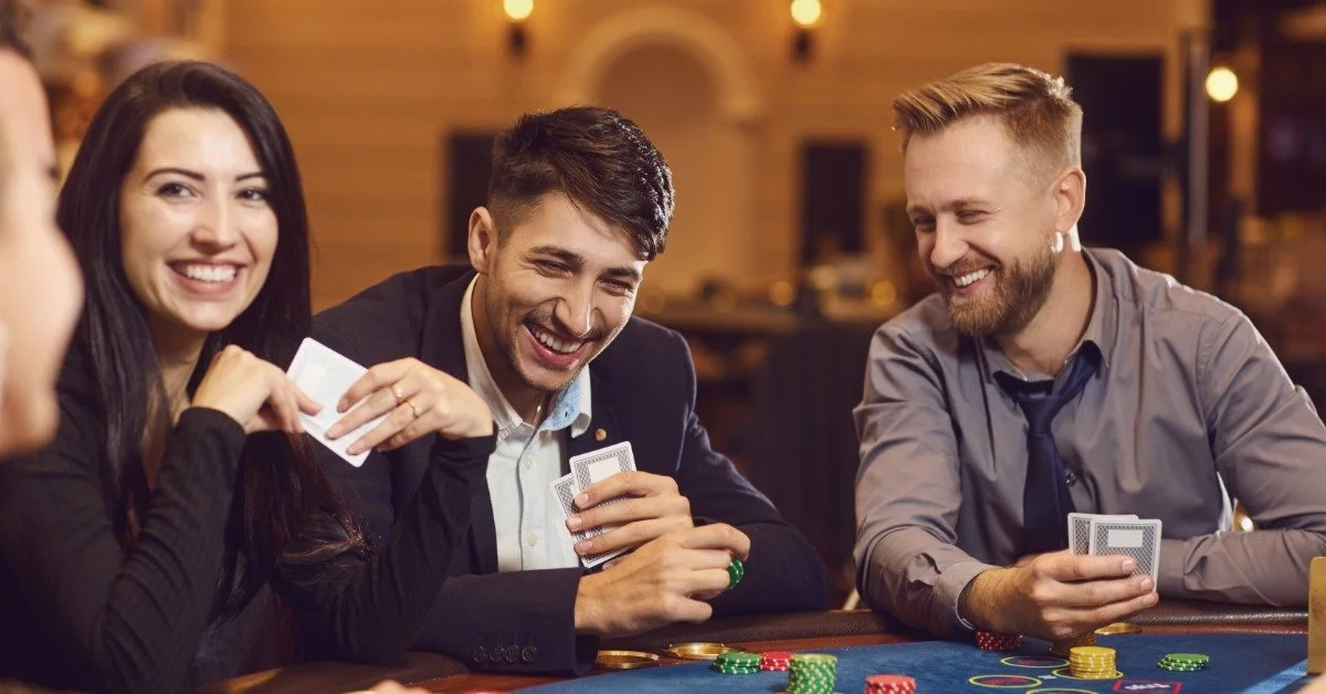 Adults at a casino table smile and laugh as they hold playing cards. Stacks of chips sit on the blue felt before them.