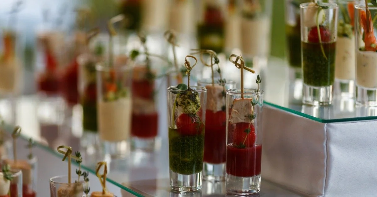 Small appetizer cups are on glass risers on a table. Each holds sauce, a cherry tomato, and mozzarella in layers.