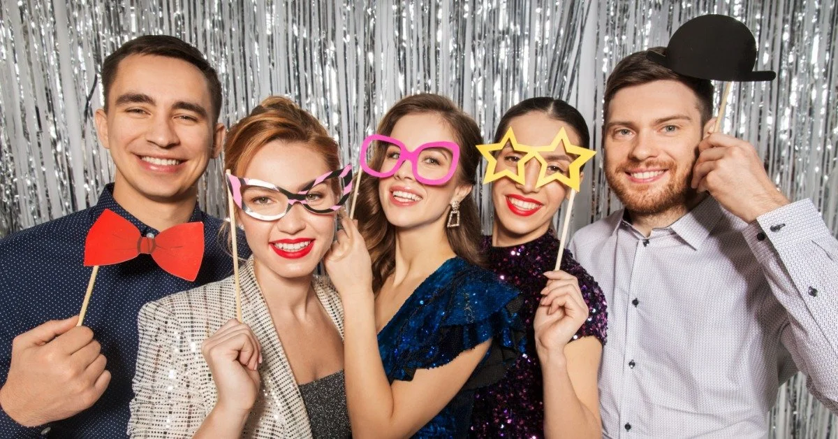 Five adults pose together in front of a silver tinsel backdrop. They hold colorful props, including oversized glasses.
