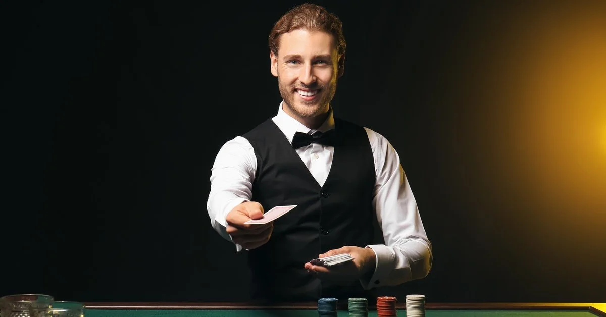A man in a vest stands behind a green gaming table and holds out a playing card. Poker chips and cards are on the table.