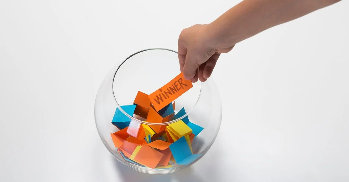 A hand holds an orange piece of paper labeled “WINNER” above a glass bowl filled with other colorful folded papers.