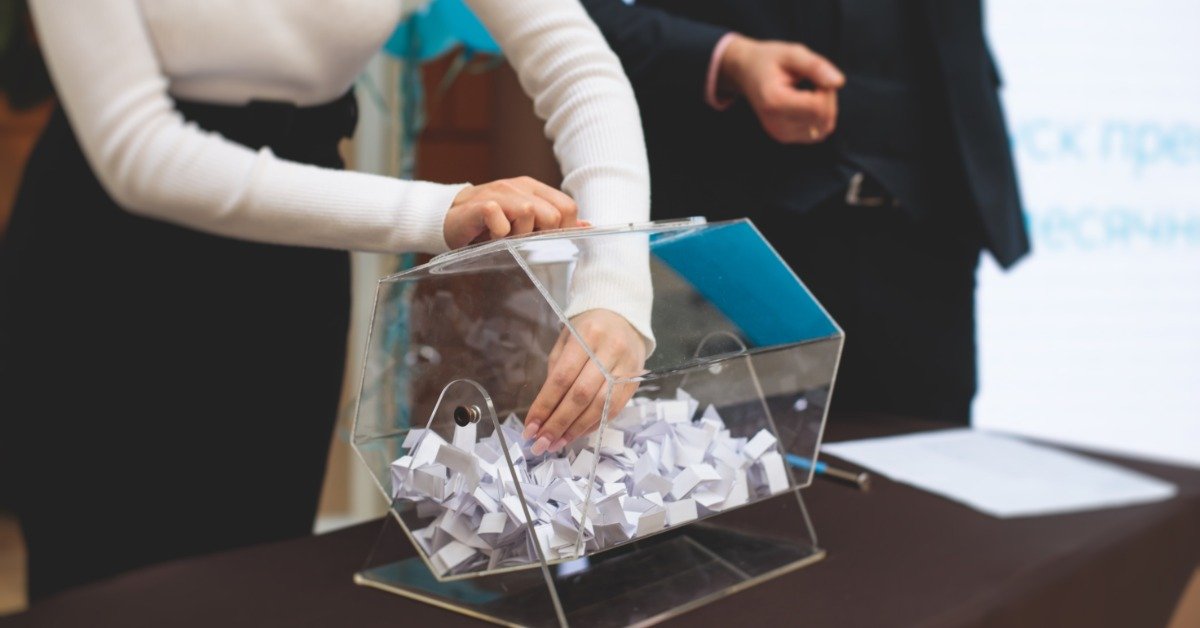 A person reaches into a clear raffle drum filled with folded paper slips. A person in a dark suit stands by the table.