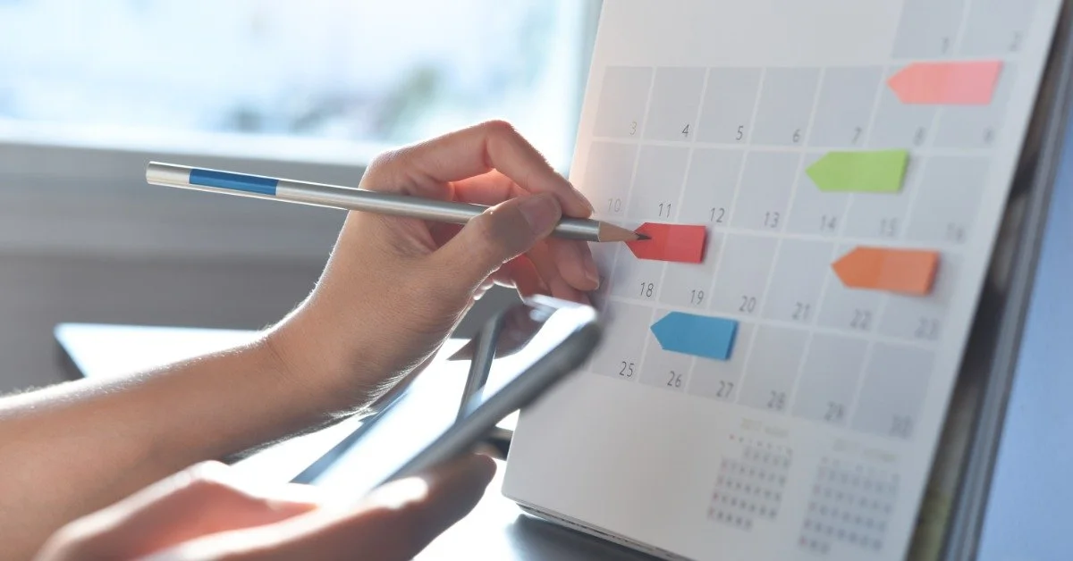 A person presses a pencil to a desk calendar covered in colorful sticky notes. They hold a smartphone in the other hand.