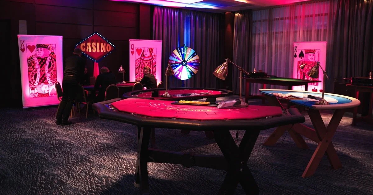 Casino gaming tables are arranged in an event space under neon lights. The room is decorated with oversized card props.