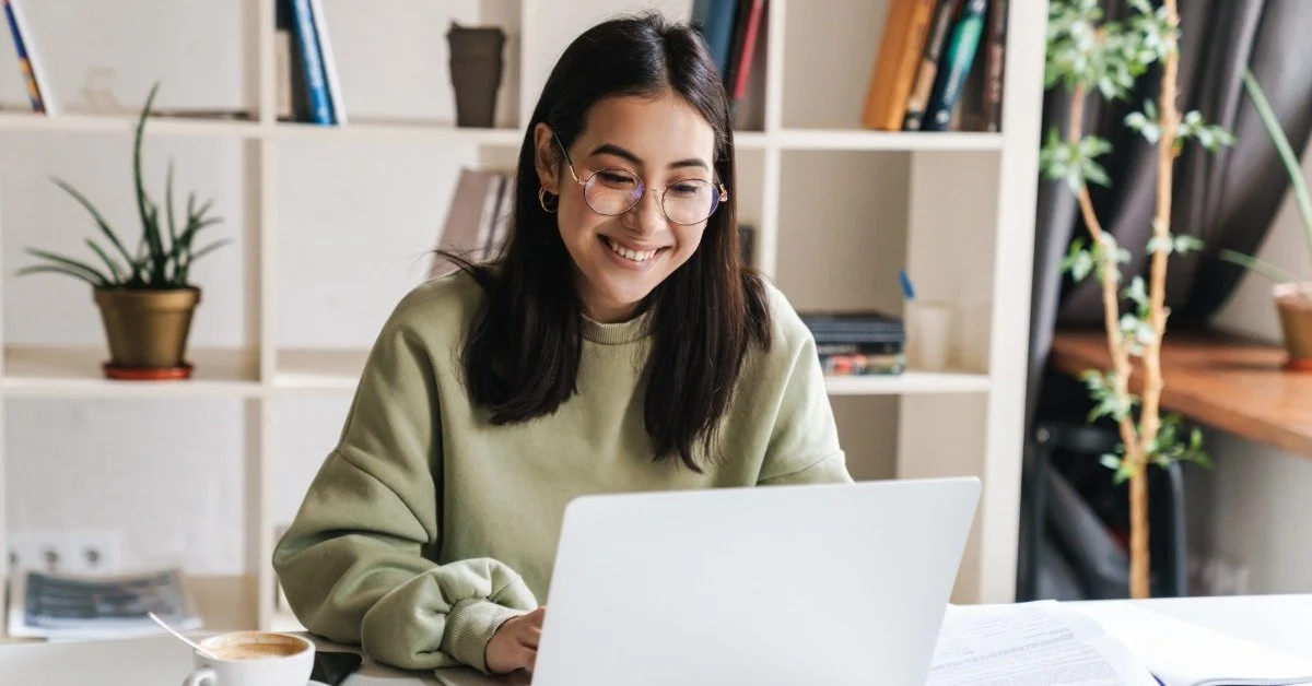 A woman works on a laptop in front of white bookshelves. Papers, colorful notebooks, and a coffee mug are on the table.