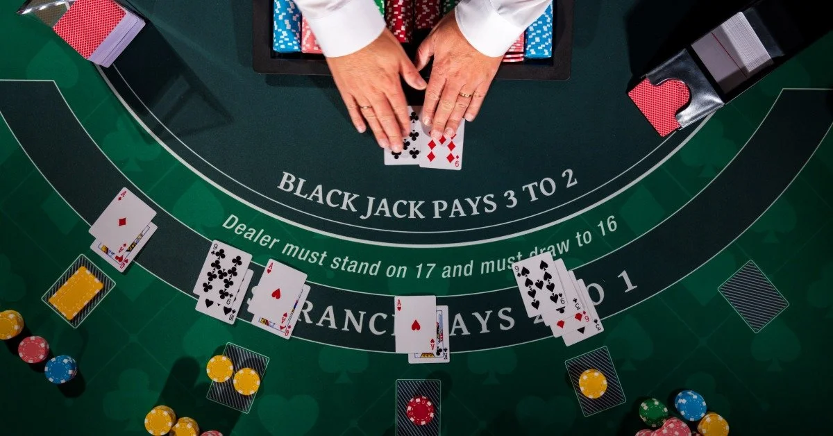 A felt blackjack table is shown from above with playing cards, colorful chips, and a dealer’s hands pushing cards.