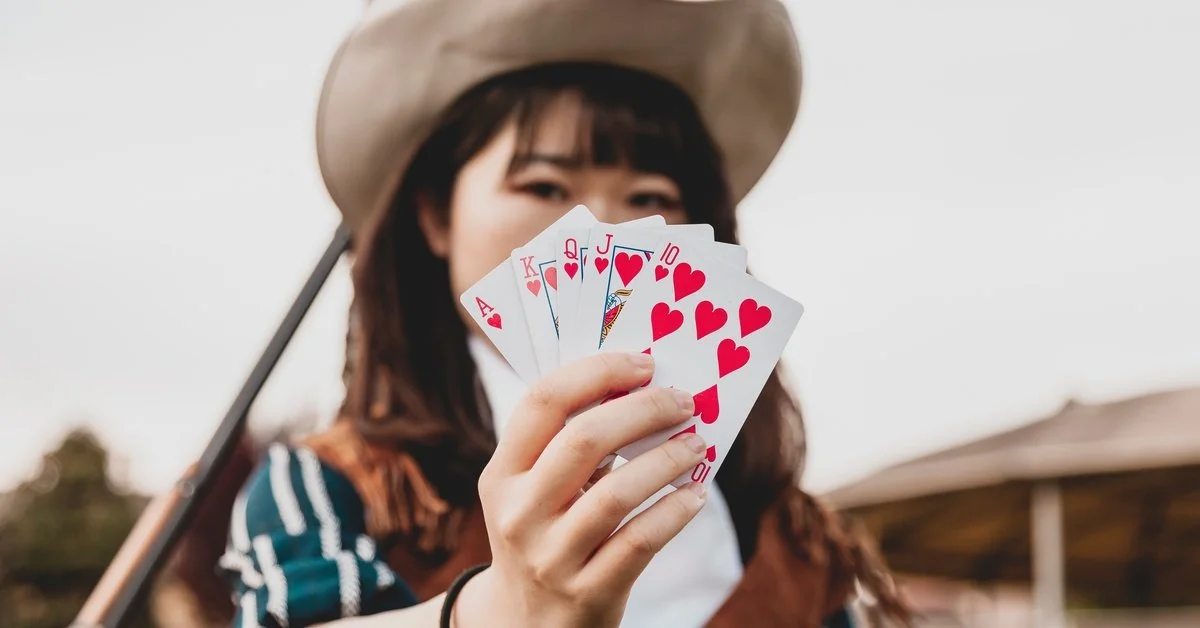 A person in a cream cowboy hat holds fanned playing cards toward the camera. The cards show a royal flush in hearts.